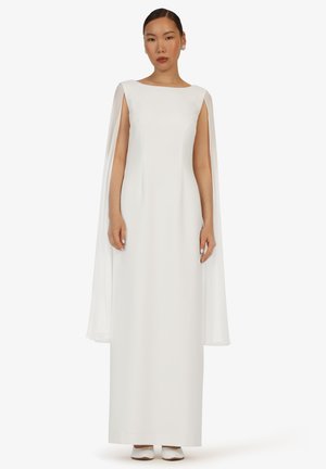 Frau trägt ein langes weißes ärmelloses Kleid mit angebrachten Umhangpaneelen und steht mit neutralem Ausdruck vor einem weißen Hintergrund.