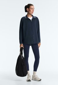 Ženska, oblečena v temnomodro zadrgo jakno, temnomodre legice, bele superge, drži veliko črno torbo, stoji pred belim ozadjem.