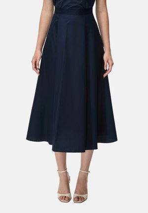 Donna con gonna midi a trapezio blu navy a vita alta, abbinata a tacchi bianchi aperti con cinturino alla caviglia, in piedi su uno sfondo semplice.