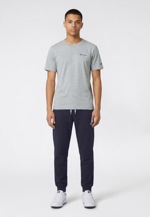 Junger Mann, der nach vorne blickend steht und ein graues Champion T-Shirt mit kurzen Ärmeln, marineblaue Jogginghosen und weiße Sneakers trägt.