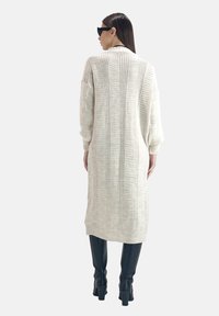Abito pullover lungo di colore crema, lavorato a maglia con texture a coste e strisce verticali, abbinato a stivali neri alti.