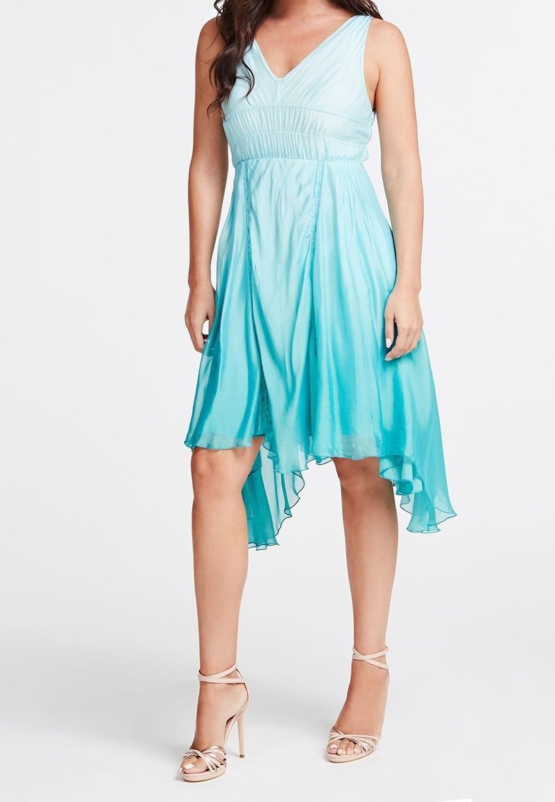 Femme portant une robe bleu clair sans manches à col en V avec un ourlet haut-bas et des sandales beiges à talons hauts à brides, debout devant un fond uni.