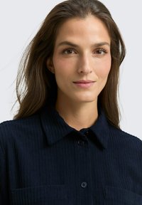 Granatowa koszula z korduroyu z kołnierzykiem, z zapięciem na guziki i dwiema kieszeniami na piersi. Charakteryzuje się prążkowaną teksturą i luźnym krojem.