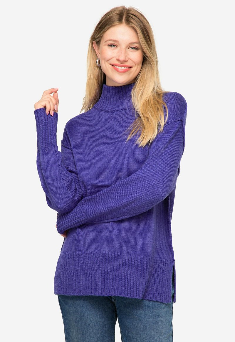 Lila gestrickter Rollkragenpullover mit gerippten Bündchen und Saum, seitlichen Schlitzen am unteren Ende und einer lockeren Passform. Getragen mit blauen Jeans.