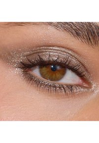 Närbild av ett öga med gyllene-brun iris, långa svarta ögonfransar och skimrande silverögonskugga mot en slät hudbakgrund.