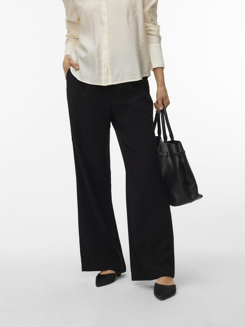 Γυναίκα που φοράει κρεμ μπλούζα και μαύρο παντελόνι με φαρδιά μπατζάκια, κρατώντας μια μαύρη τσάντα με το ένα χέρι στην τσέπη, στέκεται σε λευκό φόντο.