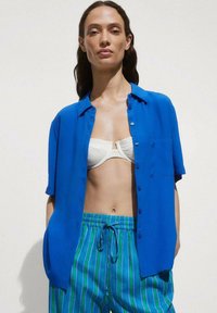 Blue short-sleeve button-up shirt with a chest pocket, paired with striped green and blue drawstring shorts. Smooth fabric textures.