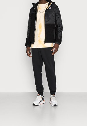 Homme portant une veste en polaire noire et en nylon par-dessus un t-shirt tie-dye orange clair, un pantalon de jogging noir et des baskets blanches et orange, debout sur un sol gris.