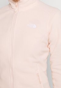 Fleece bunda světle růžové barvy, s vysokým límcem, plným zipem a bílým logem na hrudi. Hladká textura a přiléhavý design.