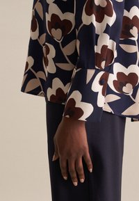 Blusa navy con grande motivo floreale in crema, marrone e rosso scuro. Presenta un polsino con bottoni e una texture liscia e setosa.
