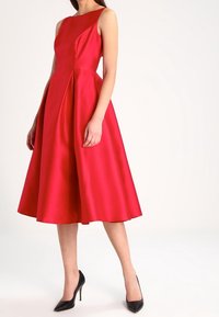 Robe en satin rouge avec un corsage ajusté, un décolleté asymétrique et une jupe évasée. Associée à des chaussures à talons hauts noires.