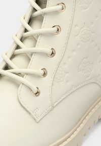 Gros plan d'une botte en cuir couleur crème avec des motifs floraux en relief, des lacets crème passant dans des œillets dorés et une couture épaisse sur la semelle.