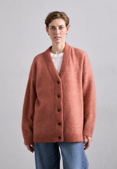 Vrouw in een losse, roestkleurige cardigan met knopen over een wit shirt en blauwe spijkerbroek, tegen een effen lichtgrijze achtergrond.