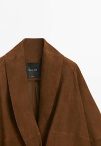 Hnědá semišová bunda se širokými klopami a černou značkou uvnitř s nápisem Massimo Dutti, zobrazena na bílém pozadí.