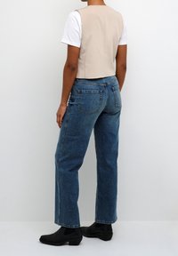 Veste courte beige portée sur un t-shirt blanc, associée à un jean en denim bleu ample. Bottines noires complètent la tenue. Design simple, sans motifs.