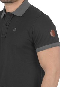 Svart polo-skjorte med grå krage, med to knapper og en liten globus-logo på brystet. Inkluderer et brunt lærdetalj på ermene.