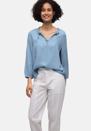 Femme souriante aux cheveux bouclés portant un chemisier bleu clair à manches longues et un pantalon blanc sur un fond uni.