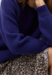 Donna con un maglione morbido di colore blu scuro e una gonna floreale con piccoli fiori viola e beige, che poggia il viso sulla mano.
