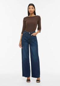 Brūns, garām piedurknēm krekls, kombinēts ar tumši ziliem, augsta vidukļa platām džinsu biksēm. Modelis stāv pārliecināti, vienu roku ieliekot kabatā, valkā melnas sandales.