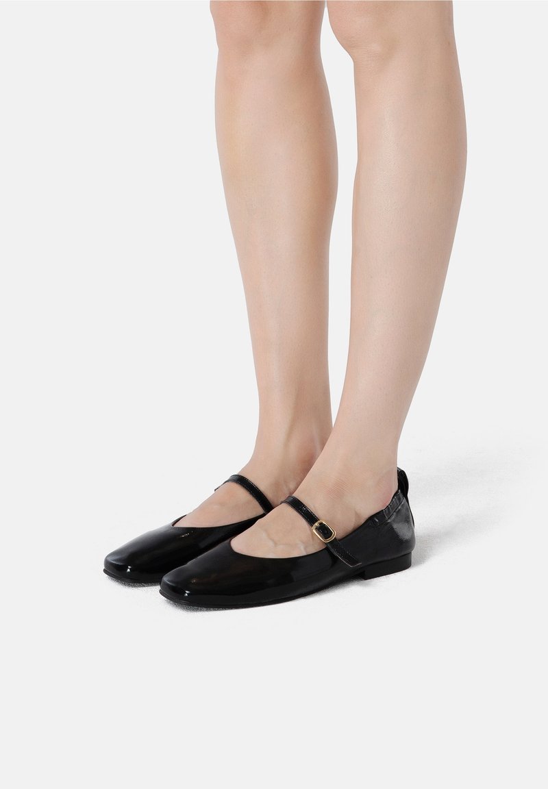Pieds portant des ballerines Mary Jane en cuir verni noir avec une petite boucle dorée sur fond blanc.