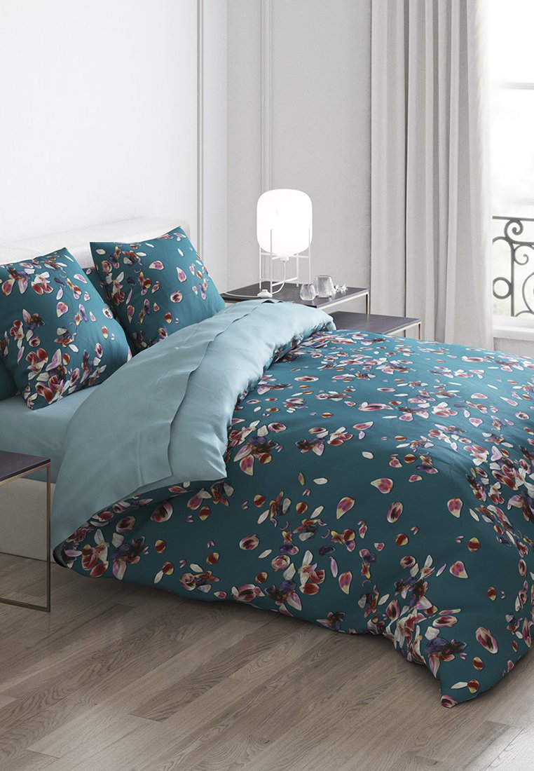 Türkisfarbenes Bettwäsche-Set mit floralen Mustern, bestehend aus zwei gemusterten Kissen und einer einfarbigen hellblauen Bettdecke auf einem Holzbettrahmen. Nachttisch mit Lampe.