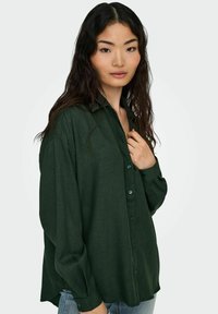 Dark green button-up shirt made of a textured fabric, featuring long sleeves, a collar, and a relaxed fit. Blue buttons accent the front.