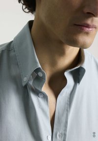Camisa a cuadros en azul claro y blanco con cuello abotonado. Presenta botones blancos y una ligera textura. Sin patrones ni acentos visibles.