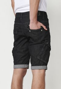 Schwarze Jeans-Shorts mit umgeschlagenem Saum, seitlicher Cargo-Tasche und zwei Gesäßtaschen. Ausgestattet mit silberner Hardware und einer strukturierten Oberfläche.