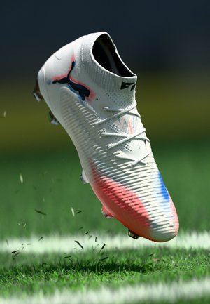 White soccer cleat with textured upper, pink and blue accents, raised sole pattern, and a low-cut ankle design, on grass with scattered blades.