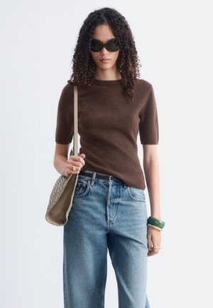 Mujer con cabello rizado que lleva gafas de sol, suéter marrón de manga corta, vaqueros azules y lleva un bolso de hombro beige con estampado.