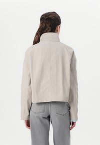 Veste beige courte avec un col haut, en tissu texturé. Présente des détails de couture et est associée à un jean gris clair.