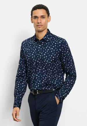 Mann trägt ein marineblaues, langärmeliges Hemd mit weißen Blumenmuster, marineblaue Hose und schwarzen Gürtel, steht mit einer Hand in der Hosentasche.