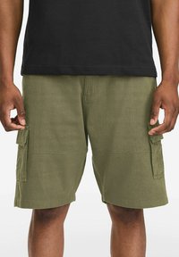 Olive green cargo shorts with two side pockets, a smooth texture, and visible stitching details, paired with a black t-shirt.