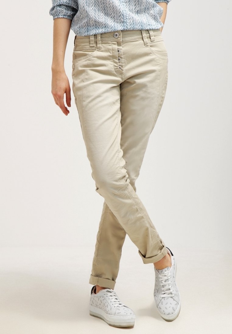 Beige puuvillachinot slim-fit leikkauksella, joissa on viisi nappilukkoa, taskut ja käännetyt lahkeet, yhdistettyinä valkoisiin kuviollisiin lenkkareihin.