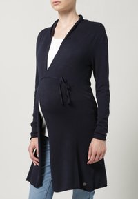 Námořnická zavinovací kardigan, měkký pletený materiál, dlouhé rukávy, detail na zavazování v pase, asymetrický lem, nošený přes bílý top, kombinovaný s džínami.