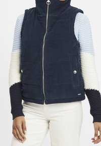 Mörkblå manchesterväst med hög krage, utrustad med dragkedja framtill, silverdetaljer och texturerade, flerfärgade stickade ärmar i blått, creme och mörkblått.