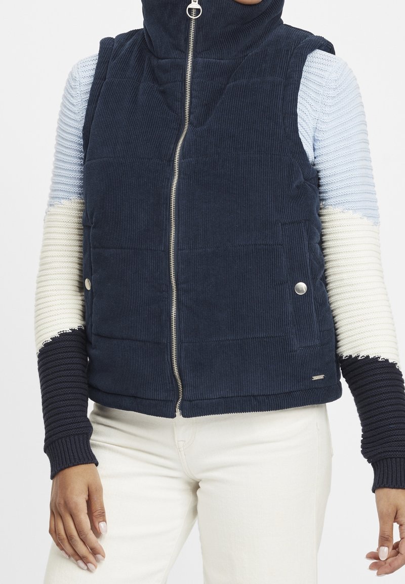 Marineblaues Cordweste mit hohem Kragen, ausgestattet mit einem Frontreißverschluss, silberner Hardware und strukturierten, mehrfarbigen Strickärmeln in Blau, Creme und Marineblau.