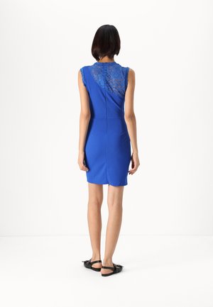 Femme portant une robe bleu roi sans manches avec un détail en dentelle sur le haut du dos, associée à des sandales plates noires, debout tournée de dos.
