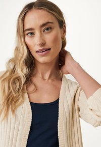 Stickad cardigan i off-white med ribbad textur och öppen framsidan, bärs över en mörkblå linne. Modellen har långt vågigt hår och delikat smycke.