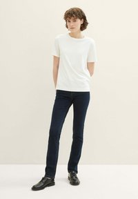 T-shirt blanche à manches courtes, coupe décontractée, associée à un jean slim bleu foncé et des chaussures noires à lacets, debout contre un fond neutre.