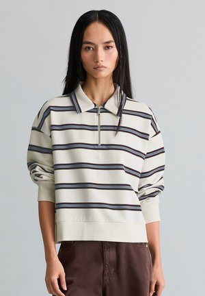 Jeune femme aux longs cheveux noirs porte un pull crème zippé avec des rayures horizontales noires et bleues et un pantalon marron sur fond gris.