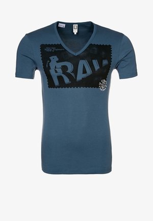 T-shirt bleu col en V avec un grand graphisme rectangulaire noir affichant le mot "RAW" et un petit texte blanc sur la poitrine.