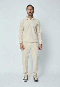 Pull-over en coton beige avec un col à revers, associé à un pantalon beige assorti. Le modèle porte des baskets noires et blanches. Design simple et ajusté.