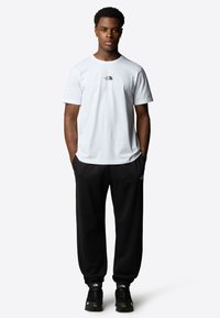 Hvid bomulds t-shirt med sort logo på brystet, parret med sorte sweatpants. Modellen bærer sorte sneakers med teksturerede såler og accenter.