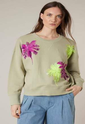 Femme portant un sweat-shirt beige avec une broderie de feuilles de palmier en néon vert et rose, associée à un pantalon plissé bleu clair, main dans la poche.