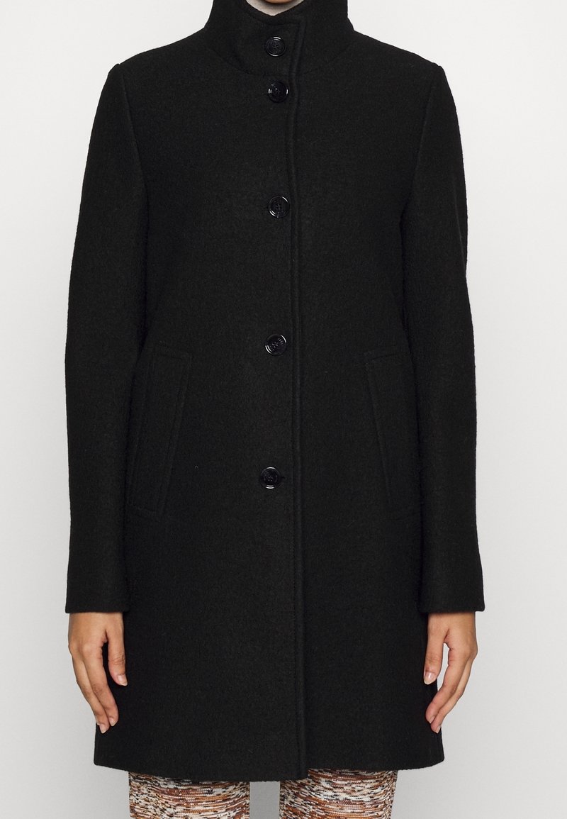 Manteau noir boutonné mi-genoux avec un col montant et deux poches latérales, porté sur un pantalon à motifs.