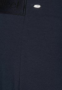 Navy blue top featuring a textured fabric, a smooth panel at the neckline, and a small metallic logo accent.