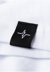 Weißer Stoff mit einem schwarzen gewebten Etikett, das ein weißes Herzschlag-Design zeigt, in die Naht genäht. Glatte Textur, minimalistischer Stil.