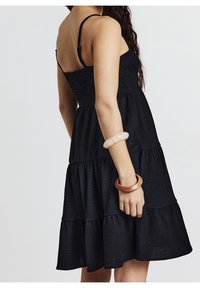 Robe noire à volants avec bretelles spaghetti réglables, tissu texturé et coupe ample ; portée avec des bracelets en bois au bras.