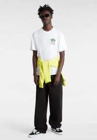 Hvid grafisk T-shirt med palmetrædesign, parret med oversized sorte bukser. En lys gul sweatshirt er bundet om livet. Sorte sneakers.